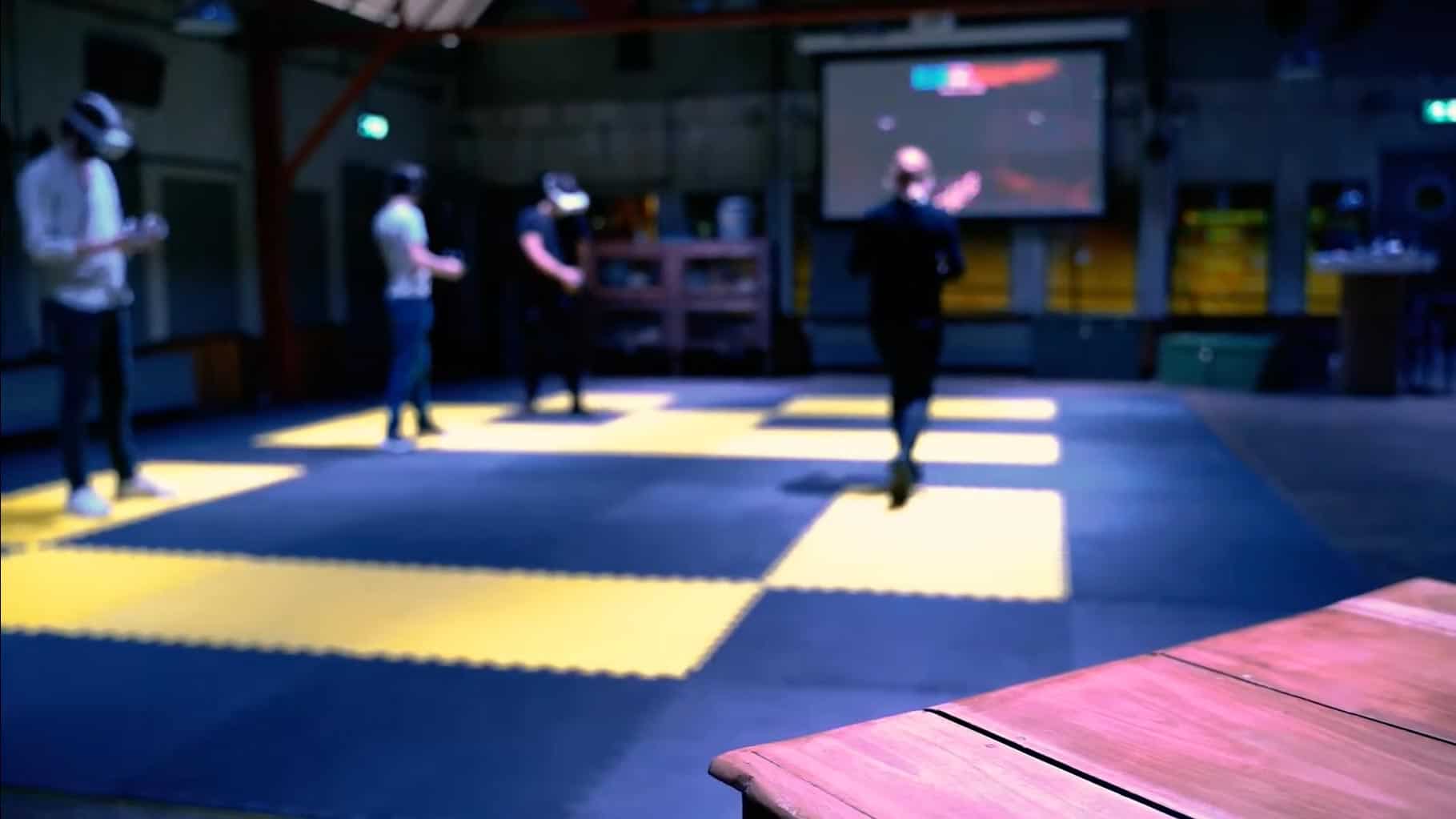 People wearing VR headsets playing a multiplayer game on a lit floor grid, with a scoreboard projected on the wall.