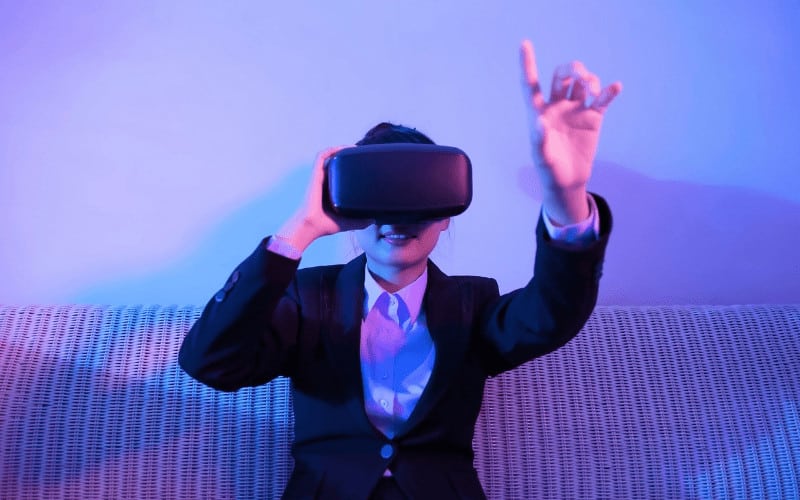 Woman in business attire wearing a VR headset with one hand raised.