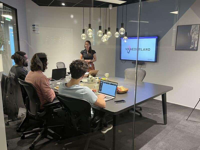 Team meeting in a glass-walled office room, with a screen showing the VR Nederland logo.
